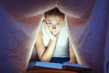 Young cheerful girl hiding under blanket and enrapt reading book