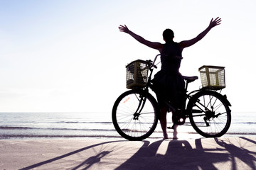 Fototapeta premium Woman silhouette with hands up spending day off bicycling on a beach against sunset sky