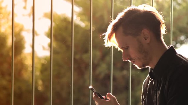 Junger Mann mit Smartphone vor einem vergitterten Fenster in warmem Licht