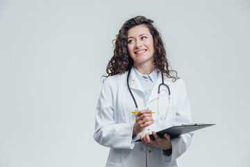 A young female doctor is on a gray background. During this, she keeps a folder with documents and thinks to write there. Displaying one of the items.