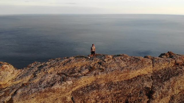 Inspiring aerial motion around fantastic bare rocky hill and person on crest top against endless blue sea at sunset