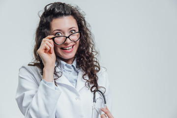 Serious young woman in a Caucasian woman in a laboratory robe, looking at you and holding the phonendoscope from his hands. Portrait of a woman in glasses with rather dark hair - a doctor wearing