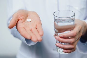 A young girl doctor takes vitamins. At the same time, she stands on a gray background dressed in a white robe. Holds a glass of water and a pill in his hands.