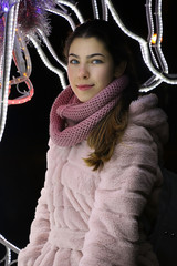 portrait of a young beautiful girl with blue eyes and dark hair in a fur coat with a scarf on the background of a garland