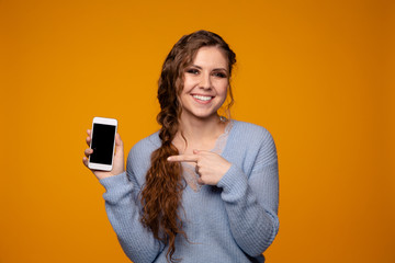 Pretty girl in a blue sweater smiles and holding her phone isolated over the yellow backgrownd,...
