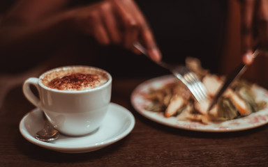 A ceramic cup of delicious hot chocolate drink on the wooden table of a dark cafe room with black female hands cutting the chicken Ceaser salad using a knife and fork in a defocused background
