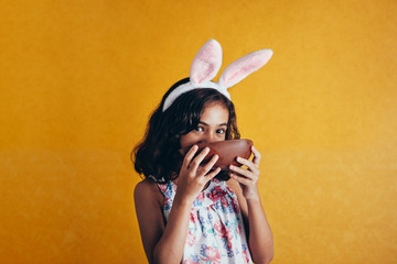 Cute little child wearing bunny ears on Easter day on color background. Girl eating chocolate easter egg