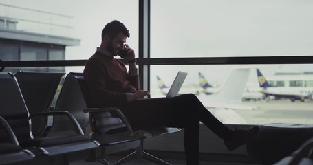 Happy with a good mood businessman working on his laptop and speaking on his phone sitting on the chair in airport , he waiting for the flight , to get boarding - Powered by Adobe