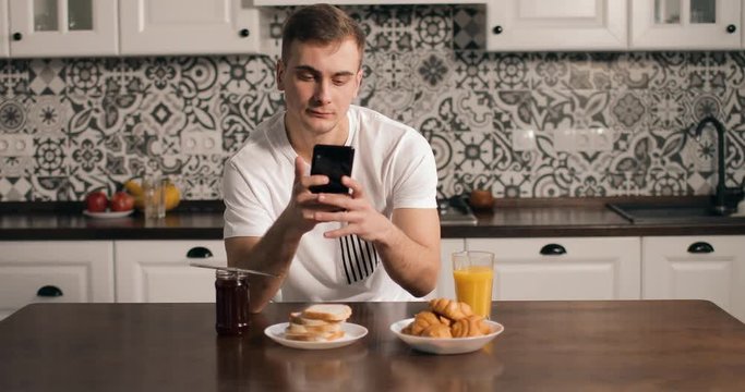 Man thinking while using smartphone  in kitchen and drinking juice in morning