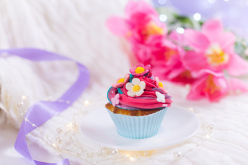 Closeup cupcake with creamy multicolored top decorated with colorful flowers with pink tulips, bokeh lights and satin ribbon on blurred background.