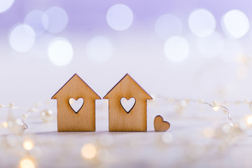 Two wooden houses with hole in form of heart surrounded by garland of white pearls on light lilac bokeh background.