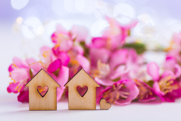 Closeup two wooden houses with hole in form of heart with tender pink cherry flowers on blurred lilac background with bokeh.
