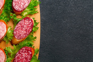Several sandwiches with sausage and salami and sauce on a black board, flat lay background with copyspace