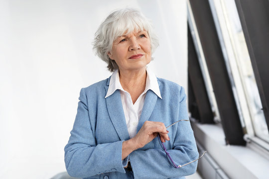 Maturity, aging and employment concept. Attractive experienced mature female entrepreneur dressed in elegant blue jacket over white shirt, pondering at her office, holding glasses and smiling