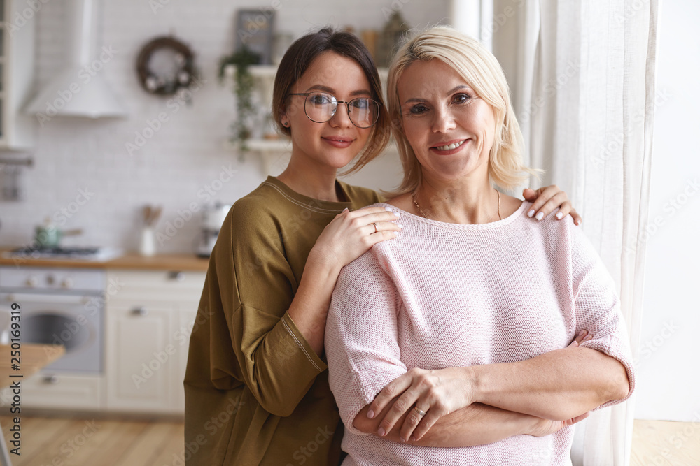 Wall mural Cozy family portrait of stylish cute teenage girl in eyeglasses embracing her charming beautiful middle aged mother. Attractive neat blonde female posing in kitchen with her young daughter