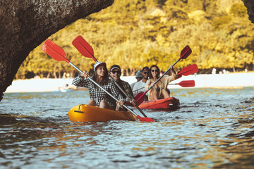Group of happy friends walks by kayaks under big rocks in the sea. Kayaking or canoeing travel photo with group of peoples