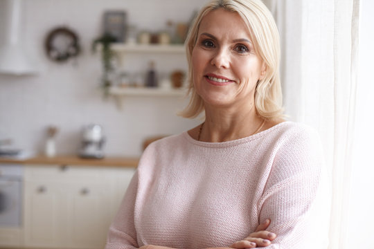 Indoor image of brown eyed middle aged Caucasian female in neat pink sweater posing against blurred kitchen interior background crossing arms on her chest and smiling joyfully. Maturity and age
