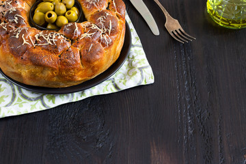 Savory pie, round monkey bread with stuffed buns oives and cheese, topped with grated cheese and black sesame bowl pitted green olives in center on black table with knife and fork olive oil copy space