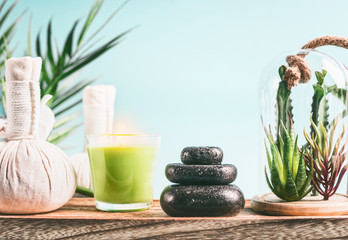 Spa concept with massage equipment:  stack of massage stones,  aromatherapy candles,  herbal stamps setting on wooden table with succulent plants at light blue background. Wellness treatment
