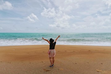 Back side view - Close up of happy traveler woman wearing black hat, t-shirt, pink short & bag raising hands with tropical sea water, waves, golden beach & white clouds blue sky background, copy space