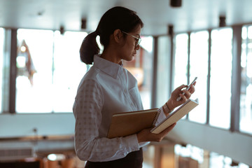 Busy Asian businesswoman wear eyeglasses using smartphone read and text message while reading a...