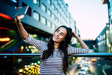 Happy smiling attractive brunette woman makes selfie on her smart phone on night city background