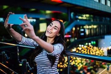 Happy smiling attractive brunette woman makes selfie on her smart phone on night city background