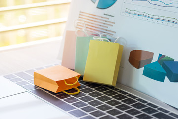 Colorful shopping bag and marketing graph report behind its on laptop keyboard. Consumer can buy product directly anywhere anytime from seller using web browser. Online shopping and e-commerce concept