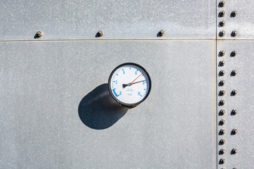 A round gauge fitted on the wall of a large galvanized steel water tank, with blue figures and two red and black needles on a white dial.