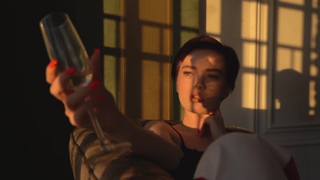 A woman with short black hair, wearing white pants and a black bra lies on a black couch and looks out the big window at the sunset. On the face of the fall sunshine, a girl holds a champagne glass 