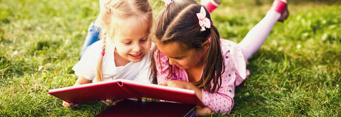 Two young girls lying on the grass outdoors reading separate books. These siblings are enjoying...