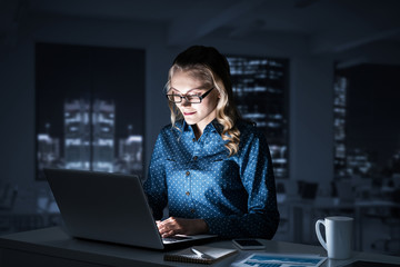 Attractive blonde working on laptop in dark office. Mixed media