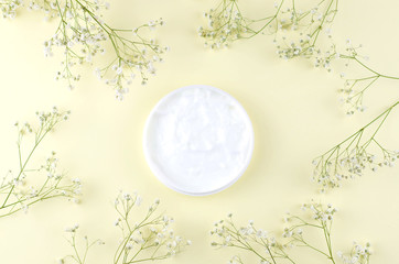A jar of cream on a yellow background with sprigs of gypsophila plant.