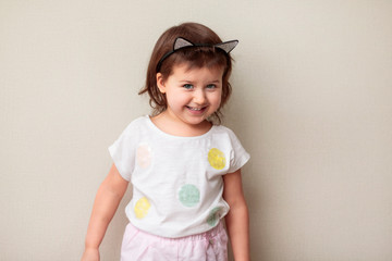 The incomplete length of a joyful little girl in a white T-shirt and pink shorts with a playful emotion is standing and pulling her arms forward, posing on a beige background, opening her mouth