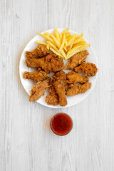 Tasty fried chicken legs, spicy wings, French fries, chicken tenders and sauce on white plate over white wooden surface, top view. Flat lay, overhead, from above.