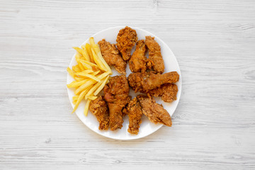 Tasty fried chicken drumsticks, spicy wings, French fries and chicken fingers on white plate over white wooden surface, top view. Flat lay, overhead, from above.