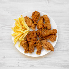 Tasty fried chicken drumsticks, spicy wings, French fries and chicken fingers on white plate over white wooden background, top view. Flat lay, overhead, from above.