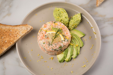 Rohes Lachstatar mit Lachs, Dill, Zitrone, Avocado und Vollkorn Toast Brot auf Porzellan Teller und Marmor Hintergrund