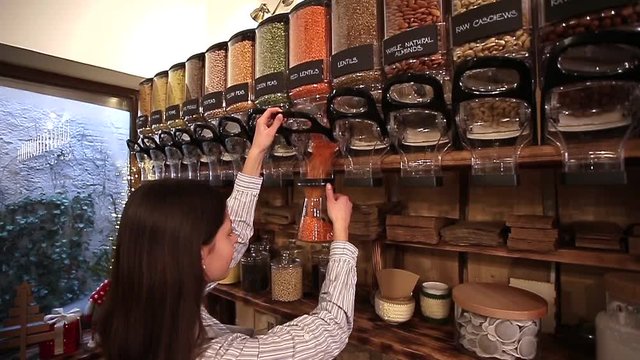 Shopkeeper working in zero waste shop. Shop assistant filling glass jar with red lentils in packaging free grocery store.