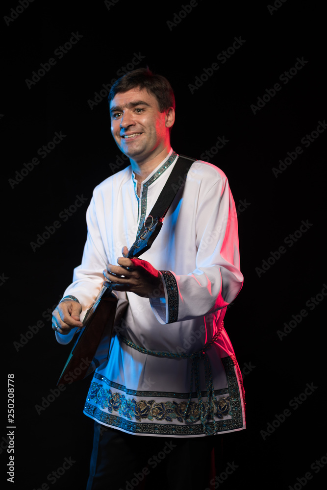 Wall mural A brunette man in a folk shirt plays a balalaika in scenic blue and red light on a black stage