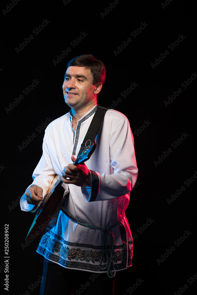 Wall mural A brunette man in a folk shirt plays a balalaika in scenic blue and red light on a black stage