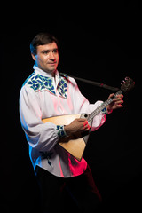 A brunette man in a folk shirt plays a balalaika in scenic blue and red light on a black stage