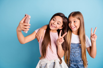 Close up photo two little she her blond brunette girls long pretty hair telephone make take selfies for mom mommy show v-sign wearing casual jeans denim t-shirts isolated blue bright background