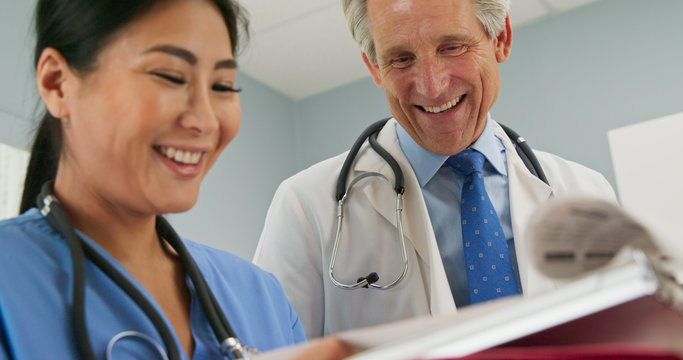 Two Happy Doctors Smiling About Good News Regarding Patient While Looking Over Paperwork Together. Senior Caucasian And Japanese Medical Professionals Working In Hospital