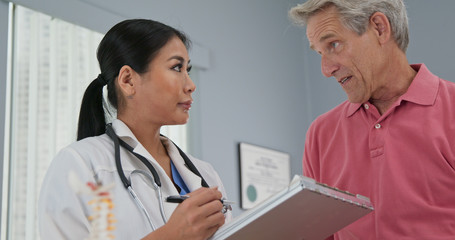 Fototapeta premium Female primary care physician taking medical history from senior Caucasian male patient. Japanese woman doctor talking to aging man and making notes