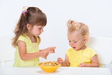Two cute little girls sisters in yellow t-shirts eating cereal flakes at the table isolated on white background