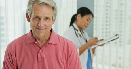 Close up portrait of senior Caucasian male patient smiling at camera while doctor works in background. Attractive older man in hospital with female physician out of focus behind him