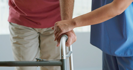 Close up on hands of physical therapist or doctor helping older male patient learn to walk with a walker. Female nurse assisting senior man with recovery after surgery