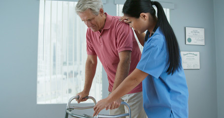 Senior Caucasian male patient learning to use walker with the assistance of Asian female physical therapist or nurse. Doctor helping older man walk again in recovery