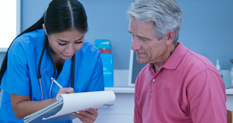 Female nurse talking to senior male patient and taking medical history. Older man sitting down in exam room while primary care doctor goes over paperwork with him